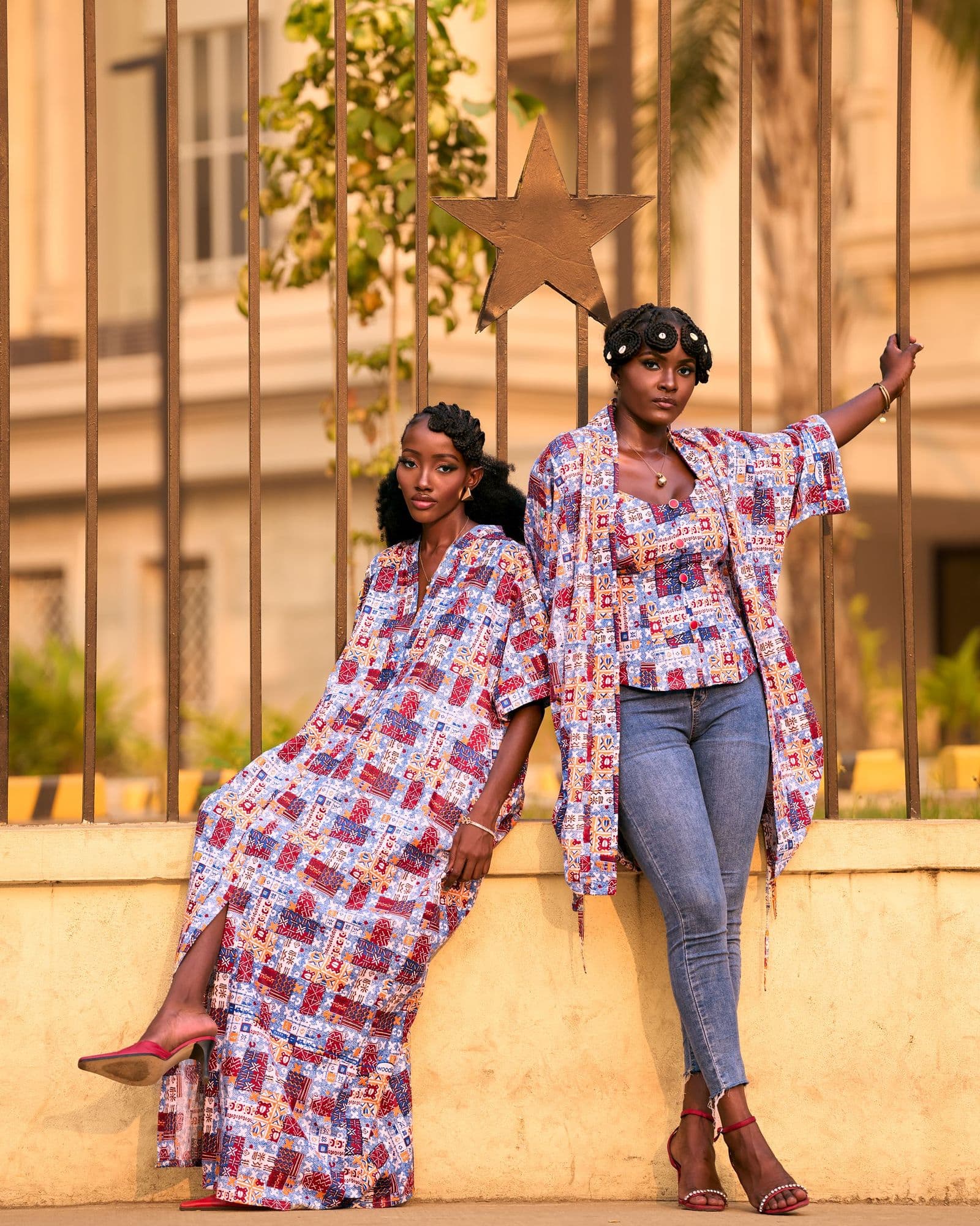 Two women wearing Zionankara wax-print pieces against an iron gate